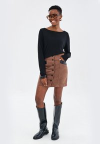 Czarny longsleeve, brązowa zamszowa spódnica z guzikami z przodu i czarnymi akcentami kieszeni, w połączeniu z wysokimi czarnymi botkami. Przezroczyste, nowoczesne okulary.