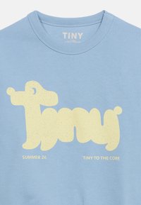 T-shirt enfant bleu clair avec un graphisme stylisé jaune d'un chien formant le mot « Tiny » et les textes « Summer 26 » et « Tiny to the core. »