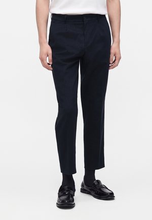 Mand iført sorte, smalle, cropped bukser, sorte sokker, sorte loafers og en hvid skjorte, står mod en ensfarvet baggrund.