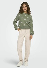 Žalias megztinis su baltais širdies raštais, derinamas su laisvomis bežinėmis kelnėmis ir baltomis sportinėmis bateliais. Minkšta tekstūra ir kasdieniškas dizainas.