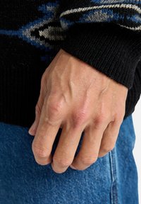 Close-up de uma mão repousando sobre denim, apresentando veias proeminentes e uma superfície texturizada, ligeiramente áspera, vestindo um suéter preto com padrões azuis e cinzas.