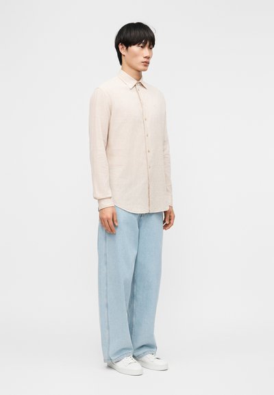 Νέος άνδρας που φορεί μια μπεζ πουκάμισο με μακριά μανίκια και κουμπιά, ανοιχτό μπλε τζιν με φαρδιά πόδια και λευκά αθλητικά παπούτσια, στέκεται μπροστά σε φόντο απλό.