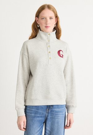 Grå fleecegenser med høy krage, tre messingknapper og et bordeaux brodert logo på venstre bryst. Skreddersydd passform med ribbede mansjetter.