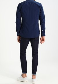 Hombre vestido con una camisa de cuadros azul oscura de manga larga, jeans oscuros remangados y zapatillas blancas, de pie de espaldas sobre un fondo blanco.