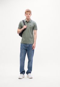 Ung mand iført en grøn polo, blå jeans, hvide sneakers og bærende på en sort skuldertaske, står mod en plain hvid baggrund.