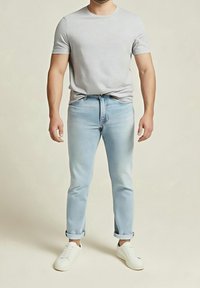 Homme portant un t-shirt gris clair à manches courtes, un jean bleu clair avec les revers roulés et des baskets blanches, debout sur un fond uni.