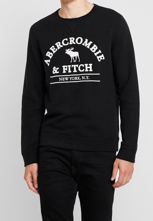 Homme portant un sweat-shirt noir avec logo blanc Abercrombie & Fitch et un graphisme élan, associé à un pantalon noir, sur fond uni.