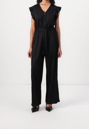 Kobieta ubrana w czarny, bezrękawnikowy kombinezon z falbanami na ramionach, zapięty na guziki dekolt w kształcie litery V, przewiązany w talii, z szerokimi nogawkami, oraz czarne, spiczaste szpilki z kokardkami.