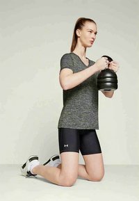 Mlada žena v sivi majici in črnih kratkih hlačah kleči in drži črni kettlebell z obema rokama na ravni prsi.