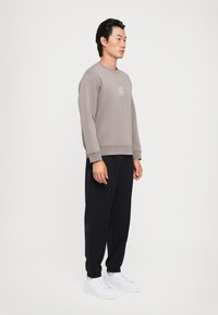 Felpa in cotone grigia con polsini a coste e logo bianco, abbinata a pantaloni sportivi neri a gamba affusolata e sneaker bianche su uno sfondo neutro.