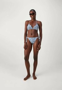 Γυναίκα που φοράει μπλε μπικίνι με σχέδιο paisley και μαύρα γυαλιά ηλίου, στέκεται ξυπόλυτη μπροστά σε απλό φωτεινό φόντο, χαμογελώντας.