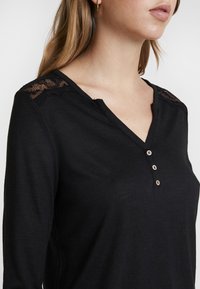 Svart långärmad topp med V-ringning, med ett spetsdetalj vid axeln och tre träknappar vid knäppningen. Mjuk, texturerad tyg.