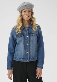 Chaqueta de mezclilla con un degradado de azul claro a oscuro, dos bolsillos en el pecho y botones de oro rosa. Acompañada de una boina de punto gris.