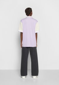 Lila Poloshirt mit kurzen cremefarbenen Ärmeln, weißen Streifenakzenten, kombiniert mit dunklen, weit geschnittenen Hosen und weißen Schuhen.