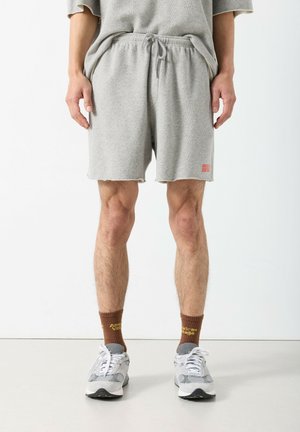 Sivé bavlnené šortky s pásom na zaväzovanie, ošúchaným lemom a malým červeným logom. Nosené s hnedými ponožkami a sivými teniskami.
