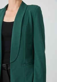 Sötétzöld blézer, hajtókás gallérral, sima anyaggal, struktúrált vállakkal, elülső zsebbel és karcsú szabással.