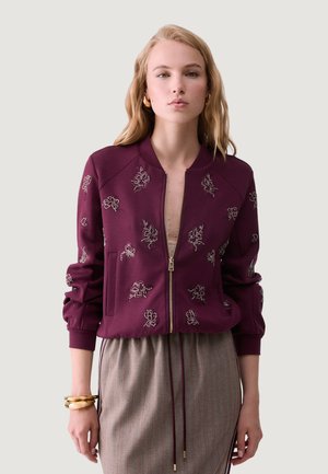 Lyshåret kvinde iført en lilla lynlåstrøje med blomsterbroderi og et beige skørt med snøre i taljen, accessoriseret med gyldne armbånd.
