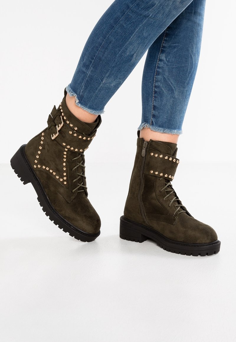 Bottes de cheville en cuir suédé vert olive avec un laçage à l'avant, une fermeture éclair sur le côté, des accents cloutés et des semelles en caoutchouc noir épais.