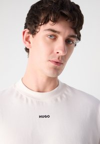 Koszulka z krótkim rękawem w kolorze kremowym, wykonana z miękkiego materiału, z prostym wzorem i małym czarnym logo "HUGO" na piersi.