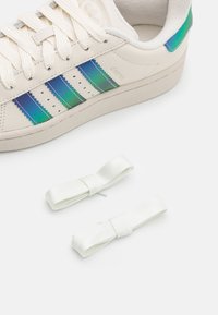 Valkoiset lenkkarit, joissa on sateenkaaren väreissä hohtavat siniset ja vihreät raidat, teksturoitu pinta, matalat nauhat ja kaksi valkoista nauhalyhtyä vieressä.