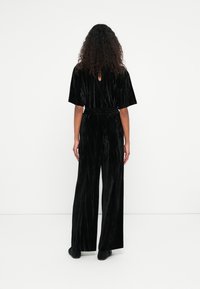 Personne aux cheveux bouclés portant une combinaison courte en velours noir avec des jambes larges, debout face à un mur blanc.