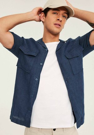 Junger Mann richtet eine beige Kappe, trägt ein offenes dunkelblaues Kurzarmhemd über einem weißen T-Shirt und hellbeige Hose, steht vor einem schlichten Hintergrund.