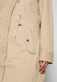 Beżowa kurtka z zamkiem błyskawicznym, wyposażona w regulowany kaptur ze ściągaczem, dwie boczne kieszenie oraz srebrny przycisk na kieszeni.