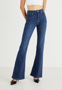 Σκούρο μπλε καμπάνα παντελόνι με ψηλή μέση, κατασκευασμένο από τζιν ύφασμα. Διαθέτει στιλ πέντε τσεπών και διακριτικές λεπτομέρειες με whisker στο μπροστινό μέρος.