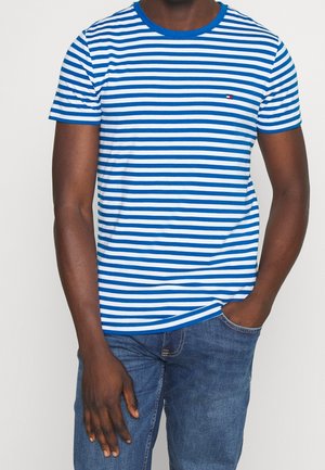Mann trägt blau-weiß gestreiftes T-Shirt mit kurzen Ärmeln und blaue Jeans, mit einer Hand in der Tasche und der anderen entspannt an der Seite.