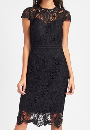 Femme portant une robe noire en dentelle mi-longue, avec des manches courtes et un empiècement en dentelle transparente au niveau du corsage et de l'ourlet.