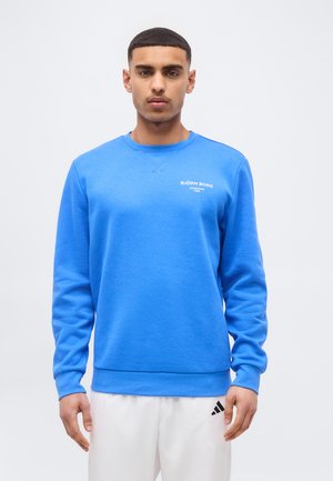 Νέος άνδρας που φοράει φωτεινή μπλε μπλούζα Björn Borg και λευκό παντελόνι Adidas, στέκεται μπροστά σε απλό φωτεινό φόντο.