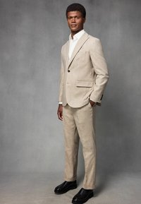 Helles beige Anzug aus strukturiertem Stoff, mit einem Ein-Knopf-Blazer, spitzen Revers und passenden schmalen Hosen. Schwarze Schuhe vervollständigen den Look.