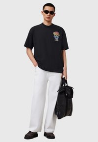 T-shirt en coton noir avec un graphique de crâne coloré, associé à un pantalon large blanc. La mannequin tient un sac noir et porte des lunettes de soleil foncées.
