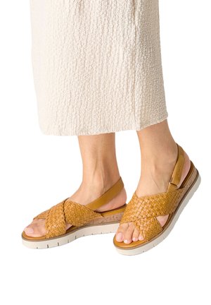 Piedi indossando sandali slingback in pelle intrecciata color cuoio con suole bianche, abbinati a una gonna texture crema.
