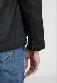 Black jacket with a smooth texture, featuring a zippered cuff detail. The hand visible is resting by the side, paired with blue jeans.