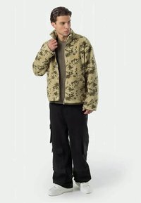 Fleecejacka i beige camouflagemönster med hög krage, tillsammans med olivgrön skjorta och svarta cargobyxor, kompletterat med vita sneakers.