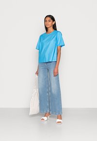 Top azul de manga curta feito de tecido suave, combinado com jeans claros de perna larga e sandálias brancas de salto, segurando uma bolsa de feira listrada.