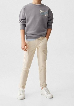 Enfant portant un sweat-shirt gris avec l'inscription "SKATE LIFE" et un motif de skateboard, pantalon cargo beige, baskets blanches, debout avec une main dans la poche.