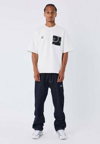 Camisa blanca de manga corta con un bolsillo negro estampado, combinada con pantalones vaqueros oscuros de corte holgado y zapatillas blancas. Elementos de diseño minimalista.