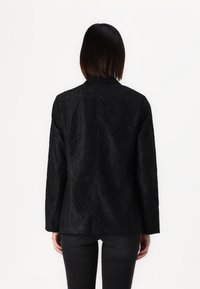 Personne aux cheveux lisses, mi-longs et foncés, portant une veste noire texturée à manches longues et un pantalon noir, debout de dos au spectateur.