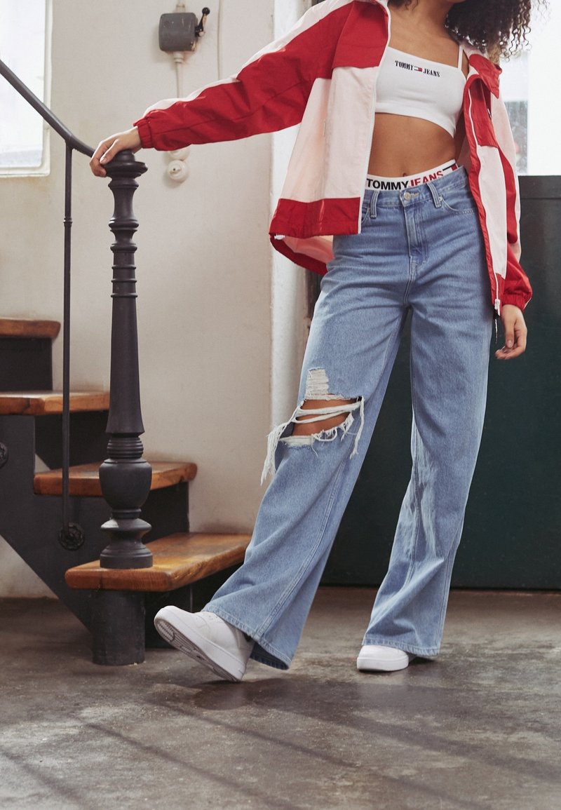 Czerwono-kremowa wiatrówka, biały top cropped z logo, jasnoniebieskie szerokie dżinsy z dużymi przetarciami i białe sneakersy na schodach.