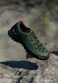 Sapatilha de trail running verde com uma superfície texturizada, sola preta e detalhes laranja. Apresenta um design com atacadores e material durável para uso ao ar livre.