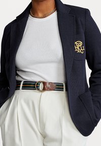 Blazer marino con un logo bordado en oro, camisa blanca de canalé y pantalones crema con pliegues, acentuados con un cinturón marrón a rayas.