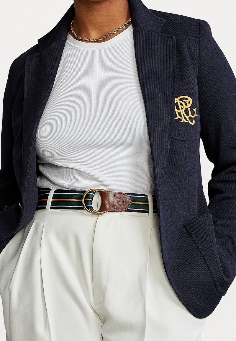 Blazer marino con un logo bordado en oro, camisa blanca de canalé y pantalones crema con pliegues, acentuados con un cinturón marrón a rayas.