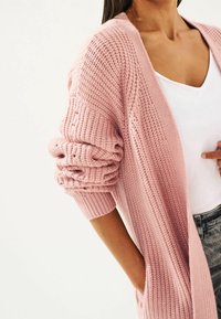 Cardigan de punto en rosa claro con textura acanalada, de frente abierto y mangas holgadas. Cuenta con dos bolsillos laterales y se lleva sobre una blusa blanca.