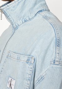 Ljusa denimjacka i blått med hög krage, frontdragkedja och en lapplåda med en logotyp. Märkbara sömnadsdetaljer.