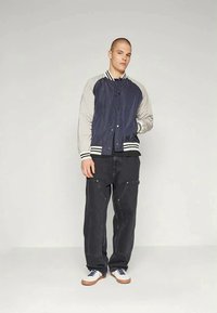Navy and gray bomber jacket with striped ribbed hem, featuring a button closure and contrasting sleeves. Model wears black cargo pants and white sneakers.