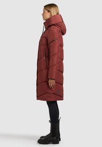 Cappotto lungo imbottito color bordeaux con collo alto, tasche con cerniera e motivo trapuntato. Abbinato a stivaletti neri alla caviglia e leggings.