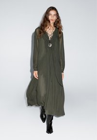 Olivgrünes, langärmliges Kleid mit Rüschendetails und V-Ausschnitt. Besitzt ein lockeres, fließendes Design und wird mit glänzenden schwarzen Stiefeln bis zum Knie kombiniert.