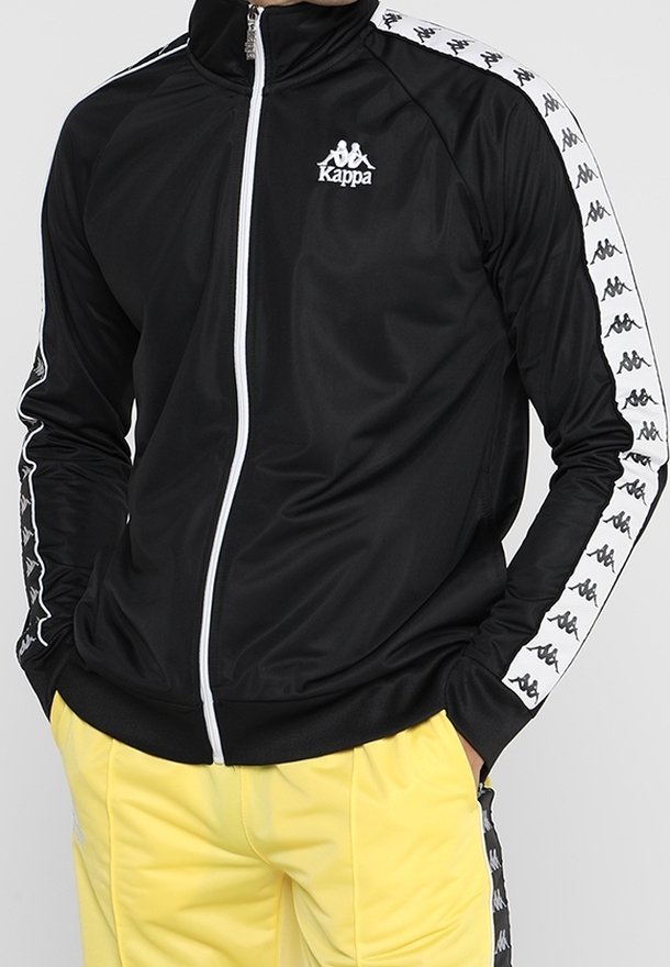 Veste de survêtement noire Kappa avec logo blanc et bandes à motifs sur les manches, portée avec un pantalon jaune, mains dans les poches, fermeture éclair avant fermée.